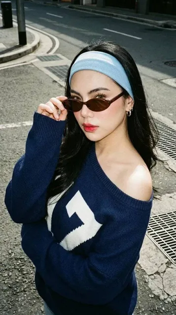 A medium close-up candid photograph of a woman named Glo on an urban street, adjusting her brown oval glasses with a serious editorial expression, wearing an oversized navy blue off-the-shoulder sweater with a white '7' graphic, in harsh natural sunlight with film grain aesthetic.