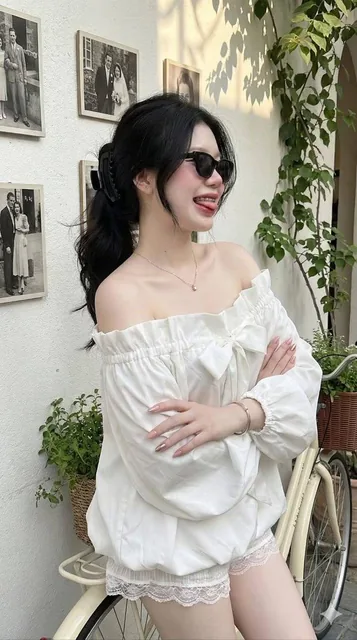 An ultra-realistic 8K mobile camera-style photo of an Asian woman with fair, radiant skin and natural makeup in a vintage European cafe setting with white plaster walls, climbing plants, and a cream bicycle. She has long black hair in a low ponytail and wears an outfit from reference image 2 with de