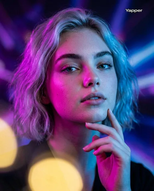 A hyper-realistic cinematic portrait of a young woman looking slightly upward with a confident, thoughtful expression. Her face is dramatically lit by neon magenta light on the left side and cool blue light on the right, set against a blurred abstract purple-blue background with soft yellow bokeh sh