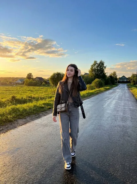 A young woman with long dark brown hair walks alone on a wet rural road during golden hour sunset, wearing a black leather jacket and gray jeans, with reflective wet asphalt and dramatic golden clouds in the background.