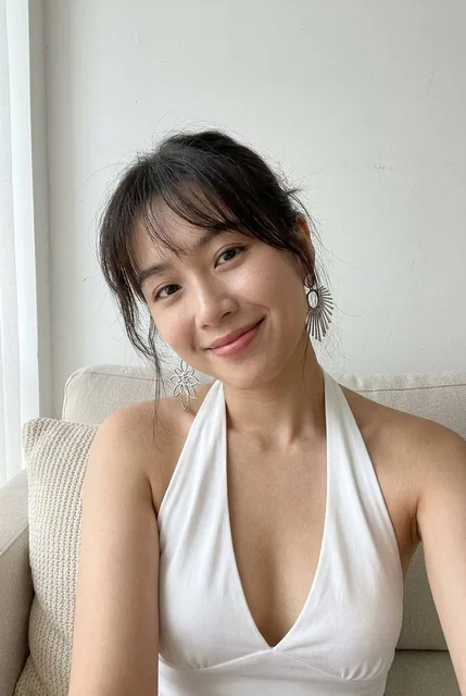 A young woman takes a close-up smartphone selfie in soft diffused daylight. She has a gentle smile, airy bangs, and dark hair, wearing a white halter top with a deep V-neck and silver statement earrings against a minimalist white background.