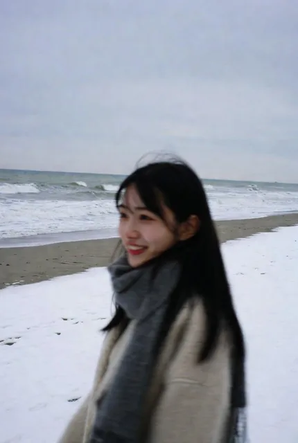 A side-profile close-up of a cute, makeup-free 18-year-old Japanese woman with long black hair, wearing a thick coat and scarf, walking alone on a snow-dusted winter beach in a cold, desolate atmosphere.