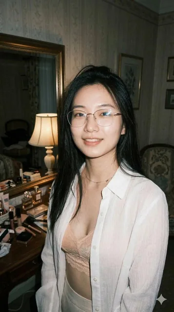 A close-up portrait of a young, petite Eastern woman with fair, translucent skin and long dark hair, wearing a fashionable semi-transparent white shirt and gold-rimmed glasses. She poses casually in front of a dressing table mirror in a dimly lit European-style room with low-saturation color tones.
