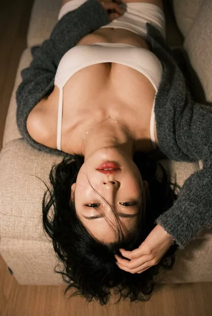 A photorealistic close-up portrait of TWICE's Myoui Mina lying back, captured from a top-down angle. She has her signature beauty marks, half-closed sultry eyes looking up, glossy lips, and wears a black camisole with a grey cardigan slipping off her shoulders.