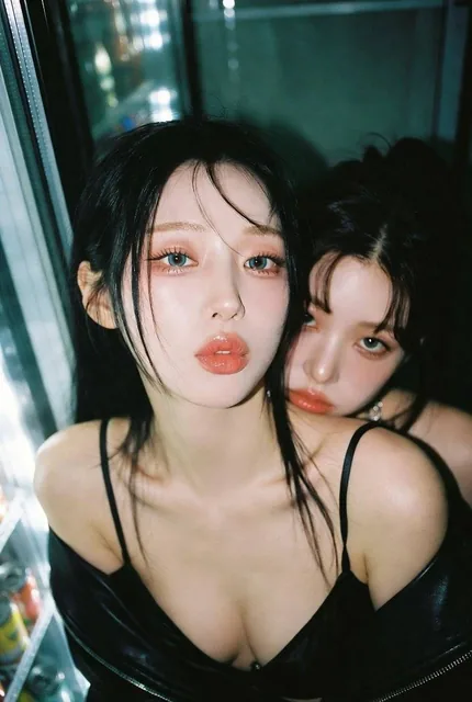Upper body portrait of two gorgeous Korean women in a convenience store at 3AM, captured with a harsh direct on-camera flash that casts hard shadows on the refrigerator glass behind them. They have flawless, luminous skin and are dressed in contrasting outfits, with intimate physical closeness sugge