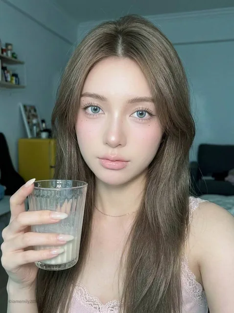 A close-up portrait of a young woman with long ash-brown hair and pale blue-gray eyes, holding a clear textured drinking glass near her face. She wears a light pink sleeveless top and has soft pink makeup, illuminated by a direct front flash in a softly blurred indoor setting.