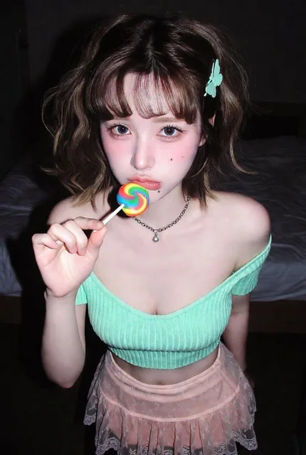 A Korean female idol posing in a dimly lit bedroom at night, photographed with an early 2000s compact digital camera with built-in flash. She wears a mint green off-shoulder crop top and nude pink mini skirt with lace trim, holding a rainbow lollipop near her mouth with an adorable pout expression.