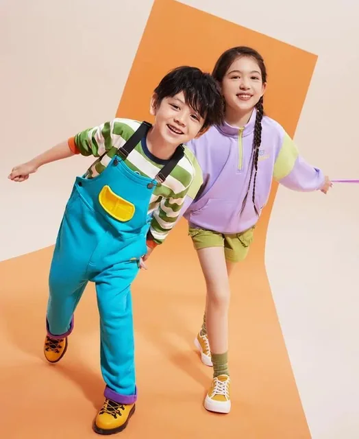 Two children walking dynamically in a minimalist studio with a diagonal orange geometric background, wearing turquoise overalls with yellow pockets, lavender and lime green hoodies, striped shirts, hiking boots and yellow sneakers, captured from a high-angle with vibrant saturated colors and soft st