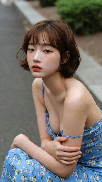 A young Asian woman with a short brown bob sits pensively on a concrete curb, hugging her knees. She wears a light blue floral sundress with a strap falling off her shoulder, under soft overcast daylight in a quiet suburban setting.