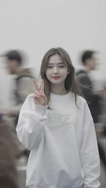 A young woman with long straight hair stands outdoors, centered in the frame, wearing an oversized white sweatshirt with a subtle logo. She smiles softly and makes a peace sign with her hand. The background features a blurred crowd in motion, with soft natural lighting, muted tones, and a cinematic