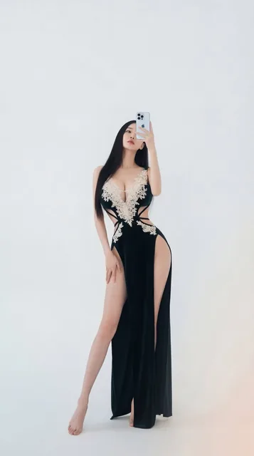 A minimalist high-fashion self-portrait of an elegant young woman with long black hair, wearing a form-fitting dark emerald green velvet gown with high slits and lace embroidery. She stands in a pure white studio, holding a phone that partially covers her face, showcasing an elongated figure and sop