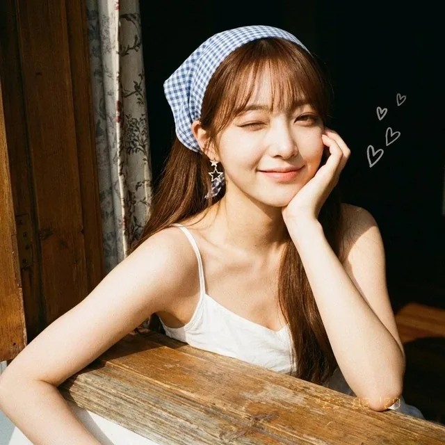 Medium close-up of a youthful East Asian woman with long brown hair and a gingham headscarf, leaning against a rustic window frame and winking flirtatiously, bathed in warm natural