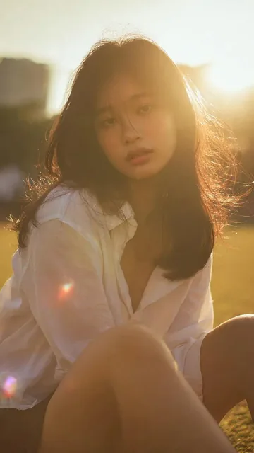 Cinematic portrait of a young Asian woman outdoors, bathed in golden hour backlight, with a soft glow and warm, dreamy atmosphere. She has a direct gaze and is wearing a white shirt and dark skirt.