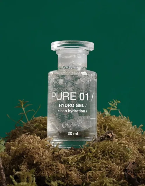 A clear glass gel bottle labeled 'PURE 01 / HYDRO GEL / clean hydration / 30 ml' rests on dense, dry moss surrounded by small natural plants and shoots. The scene features soft matte textures with no glossy moisture, set against a solid deep moss green background.