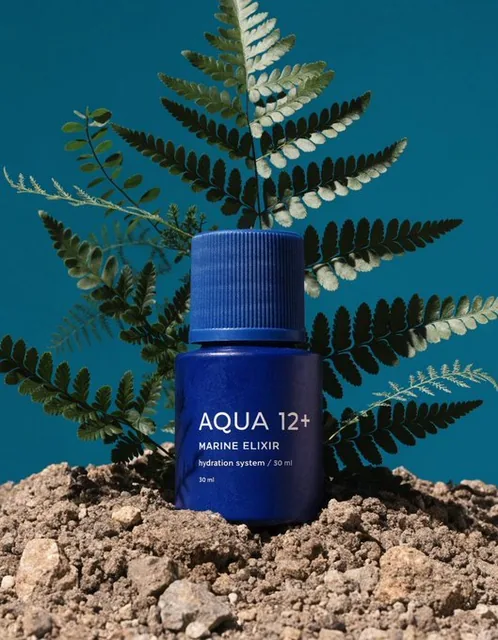 A cobalt blue serum bottle is partially embedded in dry soil, surrounded by naturally growing ferns with thin leaves gently crossing in front. The scene features matte textures with no shine or water, set against a solid deep teal background under clean natural light. The bottle label reads 'AQUA 12
