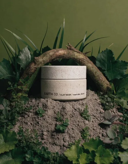 A minimalist ceramic jar is partially embedded in dry, cracked soil, surrounded by dense natural leaves and low plants. The composition is grounded with earthy lighting against a solid deep olive green background. The jar has a label that reads 'EARTH 03 / CLAY MASK / reset skin / 50 ml'.