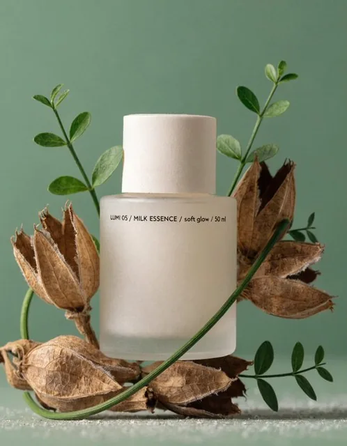 A soft milky white bottle labeled 'LUMI 05 / MILK ESSENCE / soft glow / 50 ml' is arranged among young dry shoots and fresh leaves on a solid muted sage green background, featuring matte textures and soft lighting with no water shine.