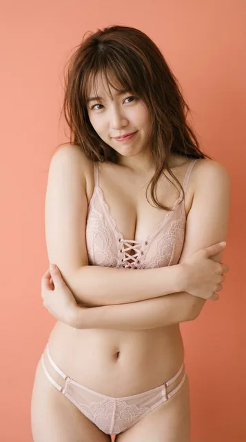 A young woman in her early 20s with chestnut brown hair, wearing pale pink sheer lace lingerie, playfully pulling her bra straps outward. Soft diffused lighting and a shallow depth of field create an intimate studio portrait.