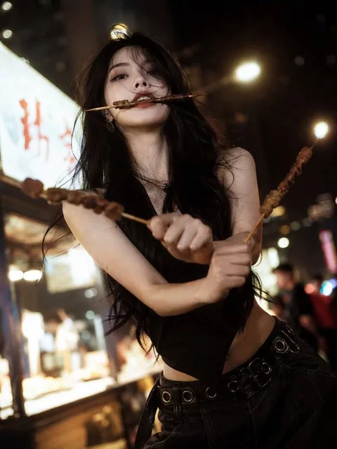 A stunning beauty with a cool, aloof expression, delicate facial features, long hair, and a slender figure. She strikes a 'three-sword style' pose with barbecue skewers: one in her mouth, one in each hand crossed. The scene is a street night market with warm yellow lighting, blurred background, film