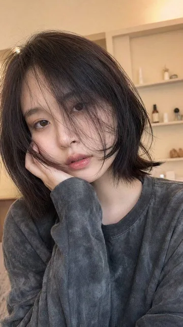 A hyper-realistic close-up portrait of a young woman with short bob hair, warm indoor lighting, natural no-makeup look, brown eyes, slightly melancholic expression, looking directly at the camera, half-body shot with tilted selfie-like angle.