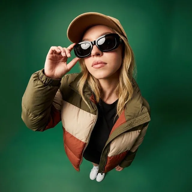 High-angle wide-angle fashion portrait of a young woman in a beige baseball cap and black oversized sunglasses, wearing a color-block puffer jacket, looking up at the camera with a confident expression in a studio with green gradient background.