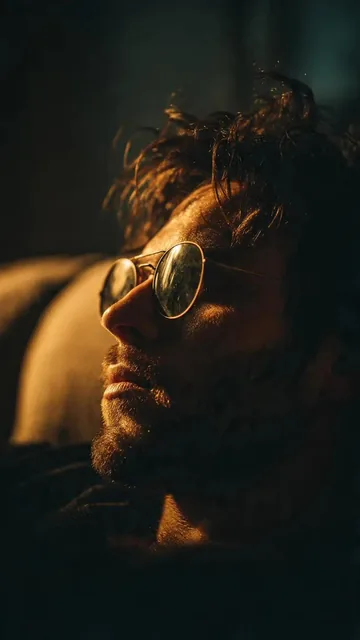 A cinematic high-resolution photograph of a man in profile wearing round-framed reflective sunglasses, seated on a soft couch with dramatic lighting highlighting his tousled dark hair.