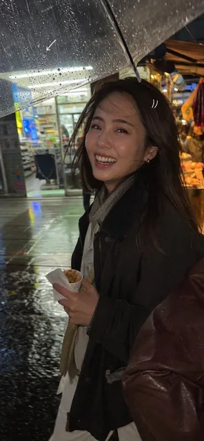 A candid vertical iPhone snapshot of an attractive Korean woman laughing mid-step in front of a rainy Seoul convenience store at night, wet pavement reflecting fluorescent light, shot through a transparent umbrella edge with raindrops and distortion, casual travel layers, natural makeup, mild motion
