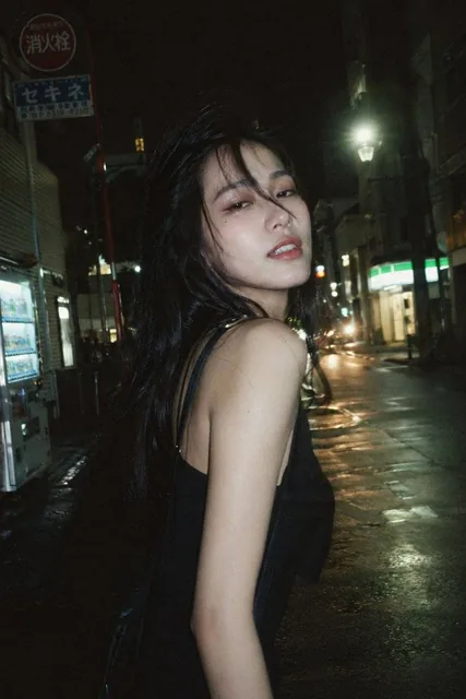 A candid night street portrait of a stunning Japanese woman walking alone on a quiet street, captured with direct flash and ambient lighting, featuring natural black hair, subtle makeup, and a relaxed expression with a hint of flirtiness.