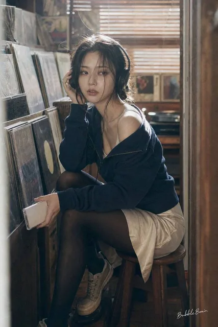 A fictional adult Korean female idol in her mid-20s sits sideways on a low wooden stool inside a tiny vinyl listening booth in a Japanese secondhand record shop. She wears a navy track jacket slightly off one shoulder, a pale cream satin slip skirt, opaque black tights, vintage sneakers, and large o