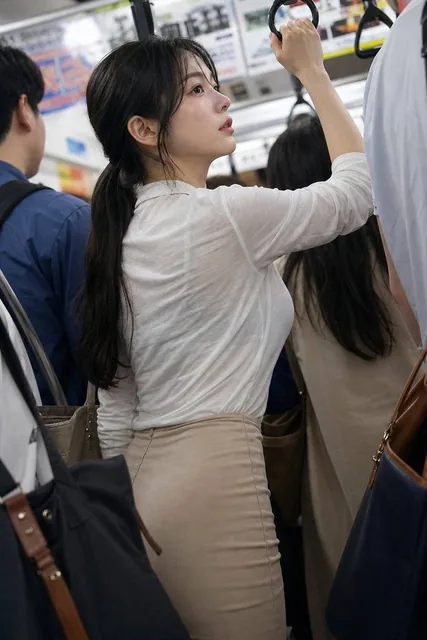 A stunning 20-year-old Japanese woman with long black hair in a low ponytail, wearing a translucent white shirt and beige tight skirt, holding a strap on a crowded morning commuter train, slightly losing balance, captured from a diagonal rear angle with motion blur.
