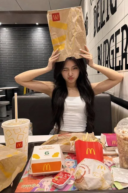 A young woman sits inside a McDonald's-style fast food restaurant, holding a brown paper bag on her head with both hands, making a playful wink and pout expression. She wears a white fitted sleeveless top with long dark hair. The table is covered with messy McDonald's food packaging including fries,
