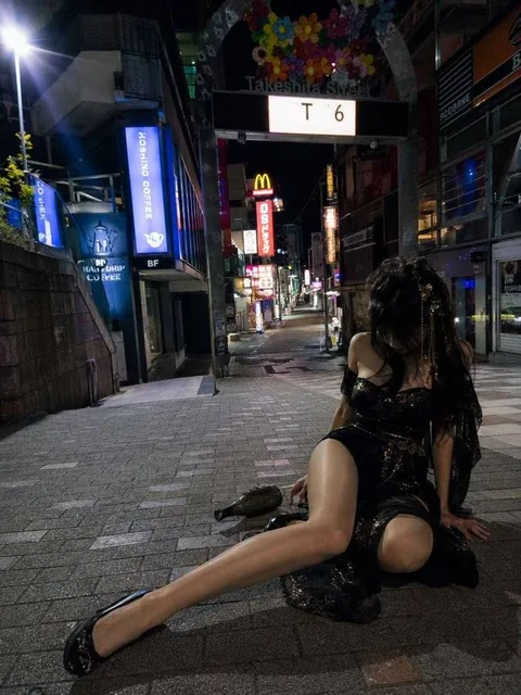 A realistic vertical night-time smartphone snapshot of a Japanese shopping street entrance, with an adult woman collapsed on the pavement in the foreground, wearing black attire and semi-transparent tights.