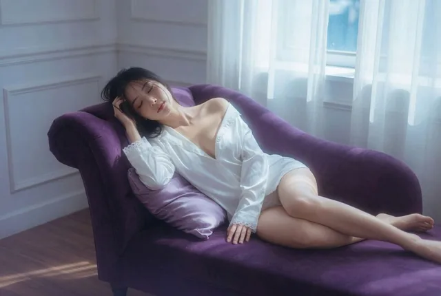 A dreamy cinematic editorial portrait of a beautiful adult East Asian woman reclining on a deep purple velvet chaise lounge beside a bright window, wearing an oversized white satin shirt with lace details, soft sleepy expression, cool daylight, overexposed highlights, film grain, shallow depth of fi