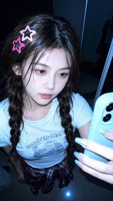 A hyper-realistic vertical smartphone selfie taken from a very high angle with a wide-angle front camera in a dark bedroom. The subject has wet messy black hair with cute glowing star-shaped hair clips, a calm melancholic expression, and is wearing a white graphic baby tee with a red plaid shirt tie