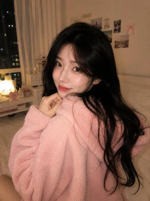 Ultra-realistic cozy bedroom portrait of a beautiful Korean woman sitting on her bed at night, turning over her shoulder with a soft shy smile, wearing an oversized fluffy pastel pink hoodie, long black hair, warm ambient lighting, city lights outside window.