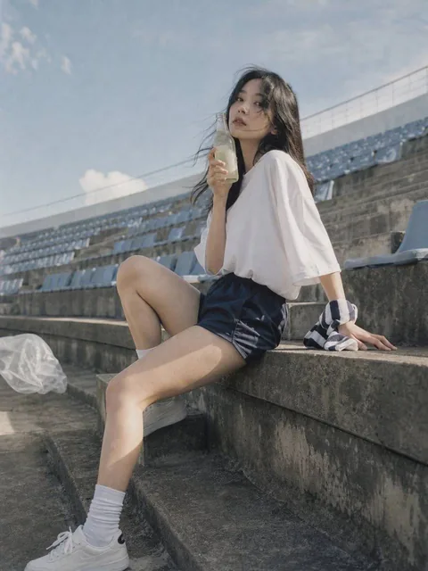 A photorealistic candid editorial portrait of an adult Korean female idol in her mid-20s sitting on empty outdoor stadium stairs after rehearsal, wearing an oversized white team-style shirt and navy shorts, with a cold glass bottle drink held near her cheek, captured in Japanese negative film aesthe