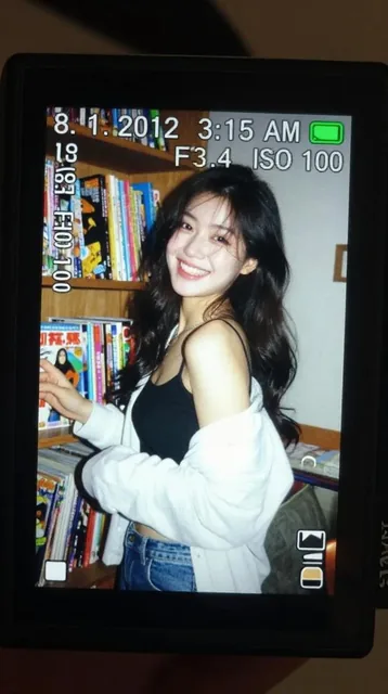 A close-up of a small digital camera LCD screen glowing in the dark, displaying a candid flash photo of a young East Asian woman with long dark wavy hair beside a comic book shelf, with visible camera UI overlays including timestamp 8.1.2012 3:15 AM and exposure data 1/30 F3.4 ISO 100.
