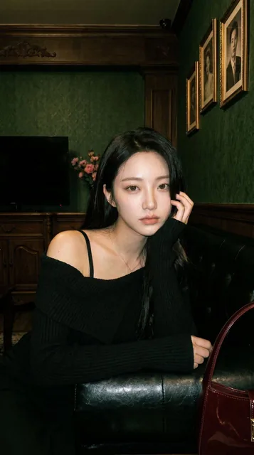 A young woman with long black hair sits on a black leather sofa in a dim vintage lounge, slightly turned away from the camera. Harsh on-camera flash creates blown highlights on her face and shoulder, while the background remains dark with green wallpaper and wooden furniture. Visible ISO noise and J