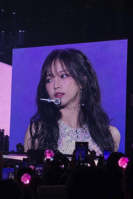A realistic concert photo taken from the audience perspective, showing a giant LED jumbotron screen displaying a K-pop idol close-up, with dark arena, silhouettes of fans, glowing pink lightsticks, and several phones recording the screen.