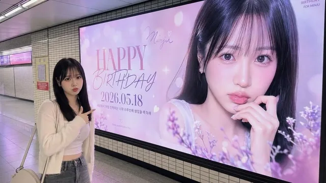 A medium-wide shot of the uploaded person standing inside a realistic Seoul subway station, proudly pointing and smiling at a massive wall-mounted LED billboard behind them displaying a fan-support advertisement featuring the same person.