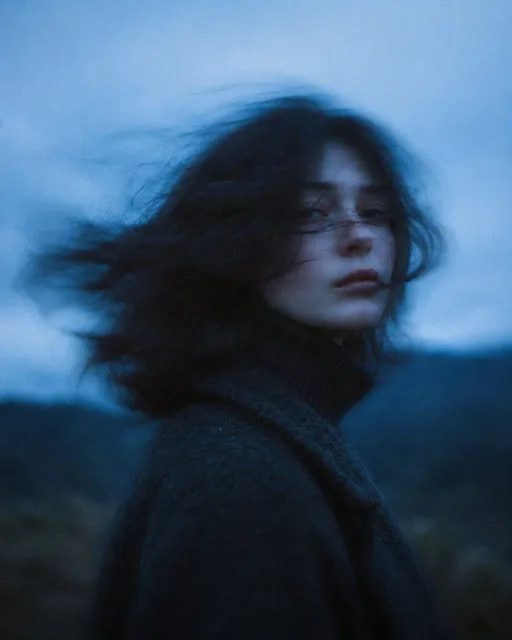 A cinematic portrait of a woman with dark wavy hair blowing in the wind, wearing a dark wool coat with turtleneck, at twilight with strong cool blue color grading, motion blur, and misty hills in the background.