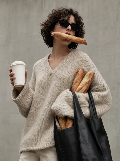 A contemporary lifestyle editorial photo of a woman with short curly hair wearing an oversized oatmeal knit sweater and sunglasses, holding multiple baguettes playfully - one in her mouth, others under her arm and in a black leather tote, with a blank takeaway coffee cup facing the camera, minimal c