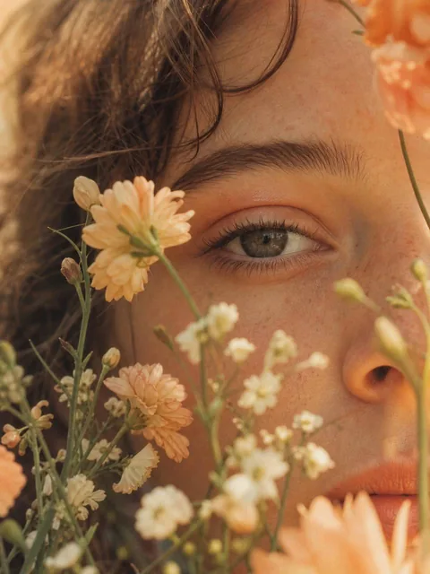 Extreme close-up photorealistic portrait of a young woman's face partially covered with delicate wildflowers, soft peach and cream blossoms touching the skin, one eye visible in focus, warm diffused sunlight, vintage film look.