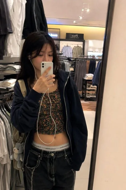 Asian girl casually walking inside a modern shopping mall, candid Y2K street fashion, low rise black flare jeans, floral spaghetti strap tank top, oversized dark zip-up hoodie, wired earphones, small shoulder bag, early 2000s aesthetic, Japanese and Korean street fashion vibe, candid iPhone snapshot