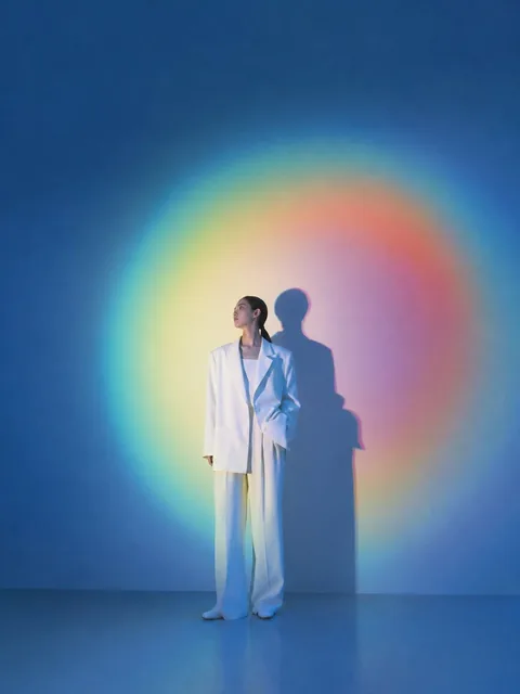 A solitary figure in oversized white clothing stands in a minimalist blue studio space, illuminated by vibrant spectral light creating a rainbow halo and elongated shadow on the wa