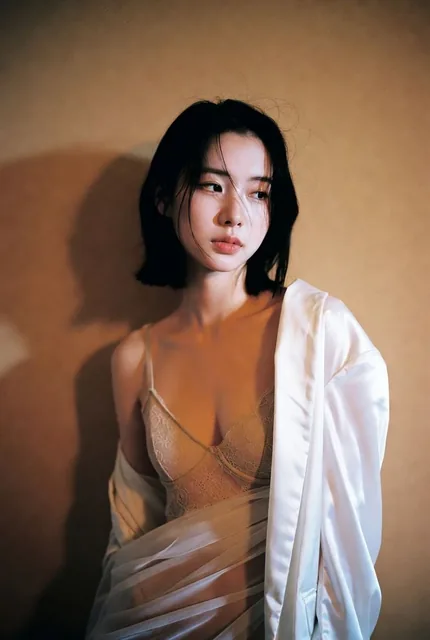 A cool-toned East Asian woman with short black hair, wearing a beige lace bra and a translucent pearl white silk shirt, leaning against a warm beige matte wall in soft indoor side-backlighting, creating a natural shadow, minimalist lingerie portrait with film texture and 8K ultra-high definition.