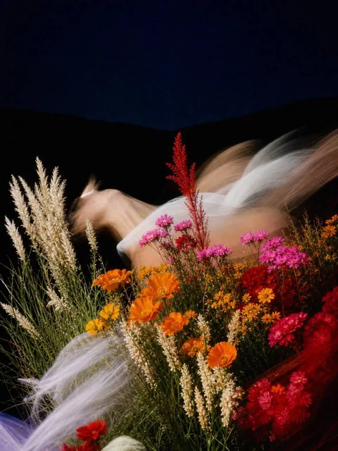 A dark surreal analog photograph of a wildflower field at night with a motion-blurred dancer, harsh flash freezing foreground flowers, deep navy sky, and rich orange, red, magenta blooms.