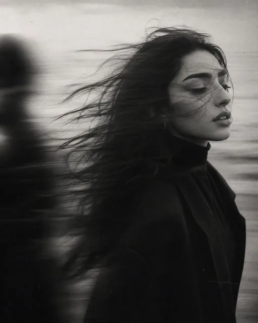 Vertical black-and-white cinematic portrait of a young Caucasian woman with sharp features and dark hair blowing in the wind, standing on a misty seashore, with a blurred male figu