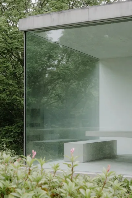 A large glass facade of a modern minimalist building reflects a dense canopy of lush green tree...