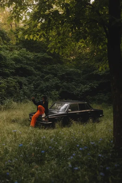 cinematic wide shot, person sitting on the hood of a vintage black boxy sedan, wearing a dark j...